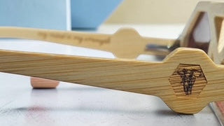 Close-up of a pair of wooden-framed sunglasses with a natural bamboo-like finish and an engraved bee logo on the temple, resting on a light surface with a blue and beige background.